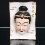 Placa devocional em porcelana chinesa, de formato retangular vertical, com bordas suavemente onduladas e filete dourado. Ao centro, cabeça de Guanyin modelada em alto-relevo, com fisionomia serena, olhos semicerrados, lóbulos auriculares alongados e cabelos reunidos em coque alto, cingidos por diadema ornamentado com motivos florais em vermelho e dourado. Policromia em preto, tons carnosos, vermelho e ouro. Campo superior à direita com inscrição vertical Báiy Guanyin, correspondente a "Guanyin de Vestes Brancas", uma das formas iconográficas da bodhisattva da compaixão; à esquerda, inscrição Shèngshn, "Montanha Sagrada". Verso com marca em azul sob o vidrado e suporte metálico para suspensão. Medidas: Alt.: 17,5 x Comp.: 12 x Larg.: 6,5 cm