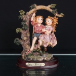 Grupo escultórico decorativo da Athena Collection, em resina policromada, representando casal de crianças sentado em balanço suspenso por cordas em galho de árvore. Menino com camisa em tonalidade avermelhada e calções azuis; menina com vestido rosa de saia ampla, ornamentada por pequenas flores aplicadas em relevo. Composição integrada a tronco arbóreo modelado, folhagens e elementos florais, sobre terreno de feição rochosa com vegetação em relevo. Assente sobre base oval em madeira, com plaqueta metálica frontal inscrita Athena Collection. Medidas: Alt.: 19 x Comp.: 14 x Larg.: 10cm.