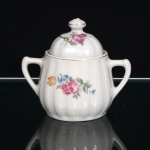 Açucareiro em porcelana nacional, esmaltagem na cor branca, de corpo bojudo molado com caneluras verticais, dotado de alças laterais estilizadas. Tampa abobadada, igualmente canelada, rematada por pomo torneado. Decoração por decalcomania policromada composta por arranjo de rosas e flores campestres em tonalidades rosa, azul e amarelo, acompanhadas de folhagem verde; friso dourado contorna a abertura, repetindo-se no remate da tampa e no pomo. Apresenta desgaste parcial do dourado e trinca vertical partindo da borda superior do corpo, vide fotos. Medidas: Alt.: 10,5 x Comp.: 12 x Larg.: 8cm.