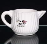 Leiteira em porcelana esmaltada branca, de corpo bojudo com caneluras verticais, assente sobre base circular, com bico vertedor curto de perfil ascendente e alça arqueada. Face frontal com decoração lúdica por decalque policromado representando vaca estilizada, com manchas castanhas, coleira com sino e laço cor-de-rosa preso à cauda, sobre vegetação. Medidas: Alt.: 10,5 x Comp.: 20 x Larg.: 12cm