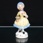 VIEIRA DE CASTRO (Rio de Janeiro, ativa aproximadamente entre 1939 e 1979). Figura decorativa em porcelana moldada e policromada, representando jovem dama trajando indumentária de inspiração vitoriana, de pé sobre base oval moldurada por volutas realçadas a dourado. Personagem com vestido de saia ampla em amarelo, acabamento inferior azul, chapéu e fita ao pescoço na mesma tonalidade, meias brancas e sapatos pretos; decote contornado por filete dourado. Na mão direita sustenta pequeno ramalhete de flores e folhagem, junto a bolsa suspensa ao antebraço. Pintura do rosto executada em tons naturalistas, com cabelos louros e detalhes fisionômicos delineados. Marca de manufatura em carimbo azul sob a base: Vieira de Castro  Rio. Apresenta desgastes na policromia e no dourado, sujidades, marcas de manuseio e sinais compatíveis com a idade. Medidas: Alt.: 14,5 x Comp.: 7,5 x Larg.: 5,5cm.