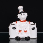 Porta-utensílios em cerâmica moldada, esmaltada e policromada, representando vaca caracterizada como cozinheira. Figura com touca de chef e avental branco pontilhado, guarnecido por gola, alças e bolsos em vermelho, patas e manchas em preto. Estrutura integrada a dois recipientes laterais abertos, destinados à acomodação de utensílios culinários. Reverso ornamentado por laço em relevo. Marca de manufatura impressa ao fundo. Apresenta craquelê no esmalte e sinais compatíveis com o uso. Medidas: Alt.: 24 x Comp.: 21,5 x Larg.: 11cm.