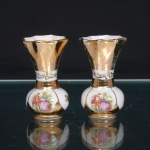 Par de vasos miniaturizados em porcelana esmaltada branca, de corpo globular, assentes sobre bases circulares, gargalo obcônico de contorno ondulado. Decoração com generosa douradura, intercalada por reservas brancas delimitadas por filetes e faixas com arabescos. Faces principais ornamentadas por estampagem policromada representando casal em cena galante, com indumentária à maneira do século XVIII, em tonalidades rosa, azul, alaranjada e verde. Faces opostas com motivos florais dourados. Medidas: Alt.: 8 x Comp.: 4,5 x Larg.: 4,5cm.