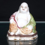 Escultura em porcelana esmaltada representando Budai, monge da tradição budista chinesa popularmente associado à prosperidade, contentamento e abundância. Figura sentada, de ventre proeminente e torso descoberto, trajando manto drapeado em verde e rosa, ornamentado por motivos florais e arabescos realçados a ouro, sobre calças em tonalidade ocre. Fisionomia delineada, com sobrancelhas, olhos e lábios destacados. Assente sobre base moldada de contorno irregular, com frisos em douradura. Medidas: Alt.: 9 x Comp.: 8 x Larg.: 7 cm.