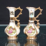 Par de pequenas jarras em porcelana esmaltada branca, de corpo piriforme, assente sobre base circular, com gargalo alto e estreito, terminado por bordo ondulado e bico vertedor pronunciado. Alças laterais vazadas, de desenho sinuoso e recortado. Superfície com acabamento iridescente, ornamentada por reservas policromadas representando cenas galantes com casal em ambiente campestre, delimitadas por filetes e campos em douradura. Numeração 85 inscrita ao fundo. Medidas: Alt.: 8,5 x Comp.: 4,5 x Larg.: 3,5cm.