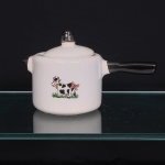 Panela em porcelana esmaltada branca, de corpo cilíndrico, com cabo lateral alongado e pequeno bico vertedor oposto, ambos esmaltados em negro. Tampa abaulada, rematada por pomo de formato arredondado com acabamento escuro. Face frontal com decoração de caráter lúdico em decalque policromado representando vaca estilizada, com manchas pretas, coleira com sino e laço cor-de-rosa preso à cauda, sobre vegetação. Superfície apresentando craquelê generalizado do esmalte. Medidas: Alt.: 14 x Comp.: 24,5 x Larg.: 13cm