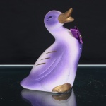 Figura decorativa em cerâmica moldada, representando pato de corpo estilizado, com pescoço recurvado e cabeça elevada. Superfície em acabamento fosco, com pintura aerografada em gradações de violeta e branco. Asas indicadas em baixo-relevo, complementadas por traços dourados alusivos à plumagem. Bico e patas em dourado; olhos delineados em negro. Aplicação de laço em fita de cetim violeta na região frontal do peito, fixado por elemento central. Base vazada, evidenciando a constituição oca da peça. Medidas: Alt.: 14 x Comp.: 12 x Larg.: 9 cm.