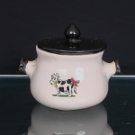 Panela em porcelana esmaltada branca, de corpo bojudo, abertura circular e duas pegas laterais curtas, com extremidades realçadas em esmalte negro. Tampa circular em porcelana esmaltada negra, rematada por pomo ogival. Face frontal com decoração de caráter lúdico em decalque policromado representando vaca estilizada, com manchas pretas, coleira amarela com sino e laço rosa preso à cauda, sobre faixa de vegetação. Medidas: Alt.: 12,5 x Comp.: 17 x Larg.: 13 cm.