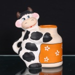 Porta-utensílios em cerâmica moldada na forma de vaca , associada a recipiente cilíndrico integrado ao dorso, destinado ao acondicionamento de utensílios de cozinha. Figura em pé com uma das patas formando alça lateral; pelagem branca marcada por manchas pretas, chifres castanhos e feições delineadas em preto, azul, rosa e vermelho. Recipiente decorado em laranja, com motivos florais brancos distribuídos pela superfície. Medidas: Alt.: 21,5 x Comp.: 20 x Larg.: 15 cm.