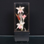 Luminária elétrica decorativa de mesa, constituída por base retangular em material sintético e redoma vertical transparente. Interior composto por arranjo de flores estilizadas, confeccionadas em tecido e fibras sintéticas, com pétalas brancas e rosadas, folhagens alongadas e elementos filamentosos translúcidos. Composição disposta sobre suporte interno, concebida para iluminação ornamental. Alimentação por cabo elétrico, com regulagem lateral. Estado conforme fotografias. Medidas: Alt.: X Comp.: x Larg.: