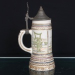 Ceramarte, Rio Negrinho - SC.(fundada em 1956). Tankard executado em cerâmica esmaltada, de corpo cilíndrico alongado, ligeiramente alargado junto à base, com alça lateral arqueada. Superfície estruturada por faixas ornamentais em relevo, compostas por entrelaçados, perlados e frisos policromados em azul, vinho e dourado. Corpo central dividido em reservas retangulares, decoradas com representações de referências arquitetônicas de Brasília, incluindo o Congresso Nacional e a Catedral Metropolitana, executadas em policromia. Tampa articulada em estanho de perfil cônico escalonado, com faixa de arabescos em relevo e mecanismo de abertura acionado junto à alça. Marca de manufatura Ceramarte  Rio Negrinho, S.C. na base. Medidas: Alt.: 22,5 x Comp.: 13 x Larg.: 9cm