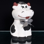 Biscoiteira em cerâmica moldada e policromada, representando vaca, com corpo utilizado como recipiente e cabeça destacável constituindo a tampa. Superfície em tonalidade branca com manchas irregulares pretas; chifres em marrom e feições delineadas em preto e vermelho. Ornamentação complementada por laços em tecido xadrez preto e branco, aplicados sobre a cabeça e ao redor do pescoço. Produção decorativa de caráter lúdico, destinada ao acondicionamento de biscoitos. Medidas: Alt.: 24,5 x Comp.: 15 x Larg.: 17cm