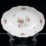 Porcelana Schmidt, Santa Catarina, Brasil (Fundada em 1945). Travessa oval em porcelana esmaltada branca, de cavidade rasa, bordo recortado e moldurado por relevos de inspiração vegetalista. Decoração em estampagem policromada, composta por ramagens floridas distribuídas pelo centro e pela aba, em tonalidades rosa, azul, verde e amarelo, com filete dourado acompanhando o contorno. Marca de manufatura Porcelana Schmidt  Mauá, Paraná, em verde, no verso. Medidas: Comp.: x Larg.:
