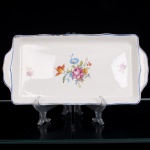 Nadir Figueiredo Ind. Com. S.A., Pedreira - SP(Fundada em 1912). Bandeja em porcelana esmaltada branca, de formato retangular, com cantos arredondados, bordo levemente ondulado e duas pegas laterais integradas. Campo central ornamentado por composição floral policromada, reunindo rosas e outras flores em tonalidades rosa, azul, amarelo e alaranjado, acompanhadas de folhagens verdes; pequenos ramos florais complementam a decoração nas extremidades. Contorno realçado por filete azul. No reverso, marca de manufatura em azul, inscrita em cartela geométrica. Medidas: Comp.: x Larg.: