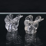 Par de figuras decorativas em vidro incolor, moldadas na forma de pássaros, com corpos arredondados, cabeças voltadas lateralmente e asas abertas. Plumagem indicada por relevos e incisões na superfície das asas e cauda. Cada exemplar apresenta pequena cavidade circular na região dorsal, destinada ao acondicionamento de vela. Medidas: Alt.: x Comp.: x Larg.: