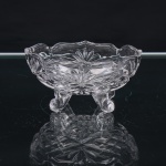 Fruteira pequena em vidro incolor prensado, de formato oval, com bojo moldado em relevos de motivos florais e geométricos, bordo recortado e ondulado. Fundo ornamentado por composição radial e paredes estruturadas em painéis moldados. Assente sobre quatro pés curvos, com terminações em voluta. Medidas: Alt.: 5,5 x Comp.: 11 x Larg.: 6 cm.