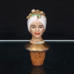 Rolha figurativa em porcelana, modelada como busto feminino. Figura com turbante branco, ornado por frutos e folhagens em relevo com douração, brincos igualmente dourados e feições realçadas por pintura policromada. Base do pescoço circundada por fileira de pontos policromos; busto com revestimento dourado. Parte inferior provida de tampão cônico em cortiça para encaixe no gargalo da garrafa. Apresenta craquelê, desgastes na douração e fissura longitudinal visível na região frontal e posterior. Medidas: Alt.: x Diam.: