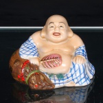 Figura de Budai executado em porcelana, representando o monge sentado, de ventre exposto e expressão sorridente, trajando manto com decoração reticulada em azul sob o vidrado. Segura leque foliforme em rosa e dourado; junto ao corpo, seu atributo iconográfico, o saco de pano (budai), modelado em marrom e ornamentado com motivos policromos, cordão vermelho e detalhes dourados. A figura de Budai, monge chinês do século X associado a Maitreya, é tradicionalmente representada com ventre proeminente e o atributo que lhe dá nome. Medidas: Alt.: 5,5 x Comp.: 9 x Larg.: 7,5 cm.