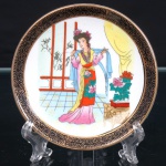 Pequeno prato decorativo em porcelana chinesa, segunda metade do século XX, decorado com composição figurativa policromada ao centro, representando figura feminina em traje tradicional chinês, portando echarpe, inserida em ambiente interior com elementos arquitetônicos, bambus e vaso com peônias. Aba delimitada por faixa de tonalidade preta com sucessão de espirais em dourado, arrematada por filetes da mesma tonalidade. No reverso, marca de fabricação impressa e Made in China Medida: Diam.: 10cm.