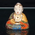 Figura de Buda executado em faiança, decoração em estilo Satsuma. Representação de Buda sentado em postura meditativa, trajando manto com decoração policromada em vermelho, azul, verde, ocre e marrom, distribuída em reservas com motivos florais e volutas, delineadas e realçadas a ouro. Face esmaltada em branco, olhos semicerrados, cabelos tratados em cachos estilizados e douração. Medidas: Alt.: 5,5 x Comp.: 5 x Larg.: 3 cm.