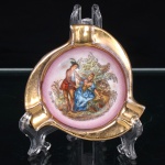 Cinzeiro executado em porcelana, formato circular com três apoios radiais destinados ao repouso de cigarros, superfície integralmente dourada e reserva central em fundo rosa. Ao centro, cena galante de inspiração pastoril, com casal em trajes europeus de evocação setecentista junto a cesto, executada em policromia por decalcomania com retoques. Reverso em porcelana branca, com vestígio de etiqueta ou marca de procedência removida. Medida: Diam.: