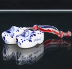 Par de tamancos miniatura em faiança, tradição de Delft, Países Baixos. Moldados segundo o klomp, calçado tradicional neerlandês, em faiança esmaltada, com decoração em azul cobalto sobre fundo branco. Faces superiores ornamentadas com reservas representando moinhos de vento, circundadas por motivos fitomórficos; laterais com ramagens e flores estilizadas. Inscrição manuscrita Holland em um dos exemplares. Peças unidas por fita têxtil nas cores vermelha, branca e azul, correspondentes à bandeira dos Países Baixos. Produção neerlandesa de caráter souvenir, vinculada à tradição decorativa da cerâmica azul e branca de Delft. Medidas: Alt.: x Comp.: x Larg.: