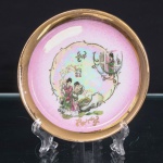 Pequeno prato decorativo em porcelana, aba em tonalidade rosa e cercadura dourada, centro com reserva de efeito iridescente, ornamentada em estampagem policromada com figuras femininas em jardim, elementos arquitetônicos, rochedos e vegetação, acompanhados de inscrição em caracteres chineses; composição secundária com figura feminina junto a estrutura arquitetônica. Filetes e arabescos em dourado. Sem marca de manufatura identificável no reverso. Medida: Diam.: