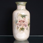 Vaso executado em faiança esmaltada, formato cilíndrico de perfil alongado, ombros arredondados, gargalo cilíndrico e bordo evertido. Superfície em tonalidade marfim, com decoração floral policroma executada à mão, composta por lírios em tons de rosa, folhagens em verde e amarelo e traços lineares em azul-escuro, gargalo ornamentado por pinceladas verticais em verde, com friso geométrico na junção dos ombros; base delimitada por filete na mesma tonalidade. Medidas: Alt.: x Diam.: