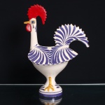 Escultura representando Galo de Barcelos em cerâmica esmaltada, assente sobre base circular. Corpo revestido de esmalte branco, com decoração pintada à mão em azul, organizada em motivos radiais sobre peito, asas e cauda; crista e barbela em vermelho, bico e patas em amarelo e olhos em azul e preto. Cauda elevada, vazada e de contorno recortado. Apresenta marca de oficina na parte inferior, de leitura não identificada. Escultura segue a tradição da olaria figurativa de Barcelos, no Minho, e tem origem na lenda do peregrino injustamente condenado, cuja inocência teria sido anunciada pelo canto de um galo assado. Ao longo do século XX, o Galo de Barcelos tornou-se um dos principais símbolos da cultura popular portuguesa. Medidas: Alt.: x Comp.: x Larg.: