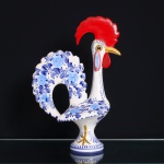 Galo de Barcelos executado em cerâmica esmaltada, Portugal, século XX. Figura zoomórfica modelada segundo a tradição do Galo de Barcelos, assente sobre base circular. Corpo revestido de esmalte branco, com decoração floral pintada à mão em diferentes tonalidades de azul, distribuída pelo peito, asas e cauda vazada; crista e barbela em vermelho, bico e detalhes das patas em amarelo. Base com marcas pintadas e numeração de oficina, parcialmente ilegíveis.Escultura insere-se na tradição da olaria figurativa de Barcelos, no Minho, e remete à lenda do peregrino injustamente condenado, cuja inocência teria sido anunciada pelo canto de um galo assado. Ao longo do século XX, o Galo de Barcelos tornou-se um dos símbolos da cultura popular portuguesa. Medidas: Alt.: x Comp.: x Larg.: