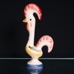 Galo de Barcelos executado em cerâmica esmaltada. Figura modelada em posição ereta, assente sobre base circular escalonada, com pescoço alongado e cauda elevada e vazada. Decoração policroma pintada à mão sobre fundo branco, com crista e cauda em vermelho, barbela na mesma tonalidade, bico e elementos ornamentais em amarelo, motivos florais e folhagens em verde e azul distribuídos pelo corpo. Base contornada por faixa azul. Sem marca de manufatura visível. Escultura insere-se na tradição da olaria figurativa de Barcelos, no Minho, associada à lenda do peregrino injustamente condenado, cuja inocência teria sido anunciada pelo canto de um galo assado. Ao longo do século XX, o Galo de Barcelos consolidou-se como símbolo da cultura popular portuguesa. Medidas: Alt.: x Comp.: x Larg.: