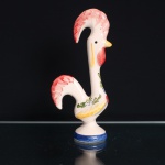 Galo de Barcelos em cerâmica esmaltada, modelado em posição ereta, assentado sobre base circular, com corpo alongado e cauda recurvada formando abertura central. Decoração pintada à mão sobre fundo marfim, com crista e cauda em vermelho, papo na mesma tonalidade, bico e elementos ornamentais em amarelo, motivos vegetalistas em verde e azul e faixa azul contornando a base. Escultura integra a tradição da olaria figurativa de Barcelos, associada à lenda do peregrino injustamente condenado, cuja inocência teria sido anunciada pelo canto de um galo assado. O motivo tornou-se um dos símbolos mais difundidos da cultura popular portuguesa. Medidas: Alt.: x Comp.: x Larg.: