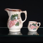 Uma(01) jarra e uma(01) caneca em faiança esmaltada de fundo marfim. Jarra de corpo bojudo, assente sobre base circular, com gargalo aberto em bico vertedor e asa lateral; acompanhada de caneca de corpo arredondado e pequena asa. Ambas apresentam decoração pintada à mão com cachos de uvas em tonalidades rosadas, acompanhados de folhas verdes e elementos gráficos em castanho. Bordas realçadas por faixa castanha aplicada a pincel. Jarra com sinais de uso, perdas pontuais de esmalte, antigo restauro junto à asa e fio de cabelo no interior. Caneca com sinais compatíveis com a idade. Medidas: Alt.: x Comp.: x Larg.: ; Alt.: x Comp.: x Larg.: