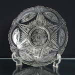Travessa circular executada em vidro moldado e prensado, de grandes dimensões, centro raso e bordo recortado. Decoração geométrica em relevo, estruturada radialmente a partir de medalhão central com estrela de seis pontas e núcleo hexagonal, circundado por campos alternados de motivos estrelados, reticulados e lanceolados. Aba ornamentada por reservas ovais e sequência de pequenas esferas em relevo. Medidas: Alt.: 5,5 x Diam.: 28,5cm.