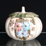 Pote com tampa executado em cerâmica esmaltada, de corpo globular e gomado, modelado em forma de abóbora. Apresenta decoração floral policromada, pintada à mão, composta por flores em tonalidades azul e rosa, acompanhadas por folhagens e elementos fitomórficos em verde, castanho e negro. Tampa conformada por folhas sobrepostas, esmaltadas em verde, rematada por haste central formando puxador. Medidas: Alt.: 16 x Diam.: 19cm.