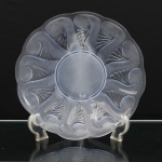 Prato circular em vidro moldado e parcialmente fosco, de bordo suavemente ondulado. Aba ornamentada em relevo por composição radial de motivos vegetalistas estilizados, alternando superfícies acetinadas e elementos transparentes; centro circular liso, delimitado por filete em relevo. Medida: Diam.: 29,5cm