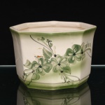 Cachepot executado em faiança, bojo de seção octogonal, perfil suavemente alargado em direção ao bordo e base moldurada. Esmaltação de tonalidade marfim, com acabamento verde no bordo e na base. Decoração floral policroma executada à mão, em gradações de verde, composta por flores, folhagens, hastes e elementos lineares, concentrada em uma das faces. Medidas: Alt.: 11 x Comp.: 16,5 x Larg.: 16cm.