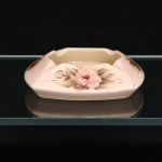 Cinzeiro de mesa em porcelana esmaltada, formato quadrangular, cantos arredondados e paredes abauladas, com cavidade central de contorno recortado e quatro apoios para cigarros dispostos na borda. Superfície externa revestida por esmalte em tonalidade rosa, com decoração floral policroma executada à mão, composta por rosas, folhagens e ramagens em pinceladas nas tonalidades rosa, verde, castanho e amarelo. Interior da cavidade sob esmalte marfim. Base rebaixada, sem esmaltação na área de apoio. Conserva etiqueta comercial fragmentária no reverso, com inscrições parcialmente legíveis. Medidas: Alt.: 4,5 x Comp.: 17 x Larg.: 17cm.