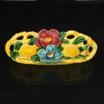 Bandeja para conjunto de sal e pimenta em cerâmica esmaltada. Japão, período Shwa, c.1930-1950. Corpo de formato oblonga, com borda vazada e duas cavidades circulares destinadas ao encaixe de saleiro e pimenteiro. Decoração moldada em relevo, composta por flores e folhagens, com esmaltação policromada em amarelo, vermelho, azul e verde. Flores aplicadas em relevo, com pétalas modeladas, acompanhadas por folhas com nervuras incisas. Reverso com marca de procedência JAPAN em verde. Apresenta craquelê do esmalte e sinais decorrentes do uso. Saleiro e pimenteiro ausentes. Medidas: Comp.: 17 x Larg.: 8cm.