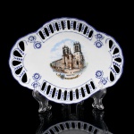 Pequena travessa decorativa em porcelana francesa, formato oval, de fundo côncavo, bordo ondulado e aba vazada, composta por sucessão de reservas oblongas e motivos vegetalistas e florais em azul-cobalto. Ao centro, representação policromada da Cathédrale Saint-Pierre de Montpellier, acompanhada do brasão municipal e das inscrições Cathédrale St Pierre e Montpellier. Decoração arquitetônica executada por processo de transferência policromada, complementada por ornamentação azul. Reverso com marca da manufatura inserida na silhueta cartográfica da França. Medidas: Alt.: x Comp.: x Larg.: