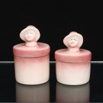 Dois(02) potes com tampa executados em cerâmica esmaltada, c.1950-1970. Corpos cilíndricos, em dois tamanhos, revestidos por esmalte em gradação de branco a rosa. Tampas circulares encimadas por figuras femininas modeladas, representadas com chapéus de abas largas e feições pintadas à mão, com olhos cerrados e lábios delineados em rosa. Decoração em tonalidades de rosa e branco, aplicada em degradê. Apresentam craquelê, perdas pontuais de esmalte e sinais decorrentes do uso. Sem marca de manufatura. Medidas: Alt.: x Diam.: ;  Alt.: x Diam.: