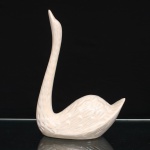 Escultura de cisne executada em cerâmica esmaltada. Figura moldada com pescoço alongado e erguido, corpo de linhas estilizadas e cauda elevada. Plumagem trabalhada em relevo, com sucessão de penas e incisões distribuídas pelas asas, corpo e pescoço. Revestimento integral por esmalte branco perolado, com variações de tonalidade decorrentes da aplicação e da modelagem. Base oval vazada, sem marca de manufatura. Medidas: Alt.: 28,5 x Comp.: 20 x Larg.: 10 cm.