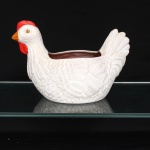 Cesta para ovos em cerâmica esmaltada, modelado em forma de galinha, com plumagem trabalhada em relevo, cauda elevada e abertura superior destinada ao acondicionamento de ovos. Revestimento predominantemente branco, com crista e barbela em vermelho, bico em tonalidade alaranjada e olhos pintados. Interior côncavo esmaltado em tonalidade castanho-avermelhada. Sem marca de manufatura aparente. Medidas: Alt.: 19 x Comp.: 31 x Larg.: 19,5cm