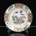 Prato em porcelana chinesa de exportação, decoração Guangcai(Cantão), família rosa, decorado em esmaltes policromos sobre o vidrado. Campo central com composição de crisântemos, peônias e outras flores, acompanhadas por aves, borboletas e ramos frutíferos. Orla circundada por reservas alternadas contendo motivos florais, frutos, pássaros e insetos, intercaladas por painéis de fundo dourado preenchidos por flores de lótus e enrolamentos vegetalistas. Paleta em rosa, verde, amarelo, azul, vermelho-ferro e preto, com aplicação de dourado e filete periférico. A organização ornamental e a paleta correspondem à tradição Guangcai, produção de porcelana decorada em Cantão destinada em grande parte ao mercado de exportação. No verso, marca estampada em vermelho com as inscrições Hand-Painted Decoration, Made in China e os caracteres Zhongguo Guangcaii, traduzidos como "Guangcai da China". Esta configuração de marca é documentada em porcelanas de exportação da segunda metade do século XX; exemplares com Hand Painted / Made in China são particularmente associados às décadas de 1960 em diante. China, República Popular da China, segunda metade do século XX, c.1960-1980. Medida: Diam.: 23cm.