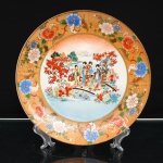 Prato de coleção em porcelana, decoração ao gosto Satsuma. Japão, período Shwa. Ornamentação em esmaltes policromos sobre o vidrado, douração e aplicações em relevo pela técnica moriage. Ao centro, cena de gênero de inspiração bijin-ga, representando figuras femininas em trajes tradicionais e criança atravessando ponte arqueada, em jardim junto a curso dágua. Peônias e arvoredo emolduram a passagem, sucedendo-se, ao fundo, pavilhões entre a vegetação e elevações montanhosas. A composição retoma o repertório iconográfico japonês de mulheres, infância, jardins e paisagens, difundido pelas estampas ukiyo-e e posteriormente incorporado às artes decorativas destinadas ao mercado de exportação. Aba com fundo ocre pontilhado em relevo, ornamentada por motivos florais, folhagens e volutas delineadas a ouro. Orla rematada por filete dourado. Reverso esmaltado em branco sem marca de manufatura. Medida: Diam.: 26cm.