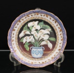 Prato de coleção executado em porcelana, esmaltagem vítrea na cor branca, composição central de natureza-morta representando arranjo de copos-de-leite (Zantedeschia aethiopica), com flores brancas e folhagem verde dispostas em vaso de duas asas, decorado em azul sobre fundo branco. O motivo destaca-se sobre campo em gradação de preto a bordô, circundado por friso de arabescos e palmetas a ouro. Aba percorrida por ramagens douradas, sucedida por faixa externa em tonalidade lilás, moldada em relevo com volutas e elementos fitomórficos. Bordo de contorno ondulado, rematado por filetação dourada. Verso sem marca de manufatura.Medida: Diam.: 25 cm.