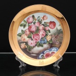 Prato de coleção em porcelana, Colección Alia, modelo Casa de Campo. Ao centro, natureza-morta floral composta por rosas em diferentes estágios de floração e pequenas flores azuis, reunidos em vaso de perfil globular com superfície craquelada; ao fundo, representação de residência campestre entre arvoredo. Em primeiro plano, fragmento de prato com motivo de rosa sobre decoração azul e branca. Aba integralmente dourada, pontuada por ornatos fitomórficos estilizados. Orla circular rematada por filete dourado. No verso, marca impressa COLECCION ALIA  Casa de Campo, e marca da manufatura Germer em baixo relevo. Medida: Diam.: 24,5 cm.