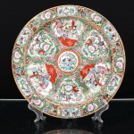 Prato de aparato em porcelana chinesa, estilo Cantão, decoração famille rose. China, século XX. Ornamentação em esmaltes policromos sobre o vidrado, com douração, distribuída em reservas contendo cenas palacianas com figuras, alternadas por composições de flores, frutos, aves e borboletas. Medalhão central com pássaros pousados sobre taça, entre ramagens floridas. Aba preenchida por ramagens, volutas e elementos fitomórficos sobre fundo dourado. Orla rematada por filete a ouro. No verso, marca apócrifa Qianlong em vermelho-ferro, com a inscrição "Qianlong nian zhi" , "Feito durante o reinado de Qianlong". Medida: Diam.: 27cm.