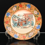 Prato de aparato executado em porcelana, decoração ao gosto Satsuma, Japão, período Shwa. Ornamentação em esmaltes policromos sobre o vidrado, douração e aplicações em relevo pela técnica moriage. Ao centro, cena de gênero representando cortejo feminino em jardim, com damas em trajes tradicionais conduzindo cavalo ajaezado, junto a pavilhão de arquitetura japonesa. Outras figuras acompanham a passagem, entre pinheiros, florações e arvoredo; ao fundo, paisagem lacustre com embarcações, casario e elevações montanhosas. A composição remete ao repertório japonês de cortejos cerimoniais com figuras femininas e montarias, recorrente no ukiyo-e e na pintura japonesa. Aba em fundo ocre pontilhado, ornamentada por peônias, folhagens e volutas, com realces a ouro. Orla rematada por filete dourado. Reverso esmaltado em branco, sem marca de manufatura aparente. Medida: Diam.: 26cm.
