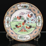 Prato de aparato em porcelana chinesa, estilo Cantão, decoração famille rose. Ornamentação em esmaltes policromos sobre o vidrado, com douração. Ao centro, composição de caráter auspicioso representando seis galos em jardim, entre formações rochosas, florações e ramos de ameixeira, acompanhados por aves em voo. Aba com fundo reticulado em vermelho-ferro, compartimentada por quatro reservas contendo pássaros e motivos florais; friso externo preenchido por flores, frutos e folhagens sobre fundo dourado. Bordo contornado por filete a ouro. No verso, marca apócrifa Qianlong em vermelho-ferro, com a inscrição "Qianlong nian zhi", "Feito durante o reinado de Qianlong", alusiva ao reinado de Qianlong (17361795), dinastia Qing. Medida: Diam.: 27cm.